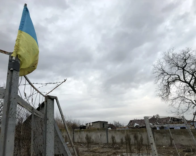 Український прапор на іржавій брамі з колючим дротом біля зруйнованих будівель
