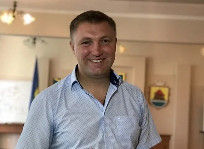Посміхнений чоловік у блакитній сорочці в кабінеті з прапором і гербом