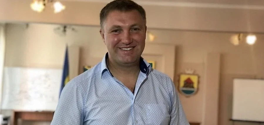 Посміхнений чоловік у блакитній сорочці в кабінеті з прапором і гербом