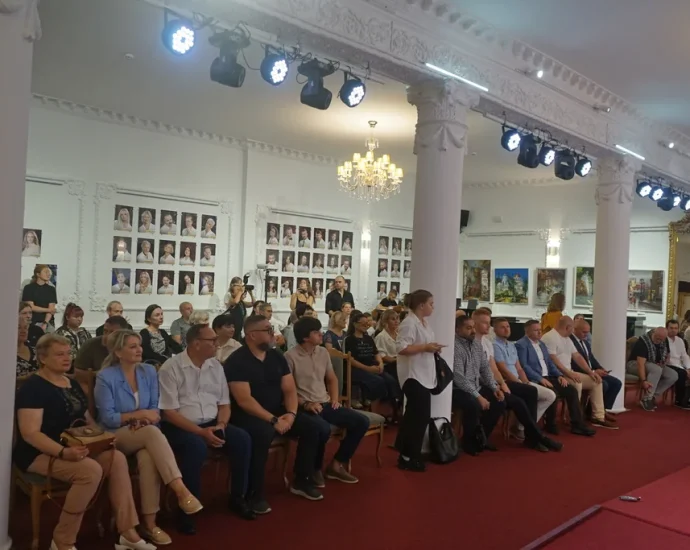Глядачі сидять у залі театру з колонами, люстрою та портретами на стіні