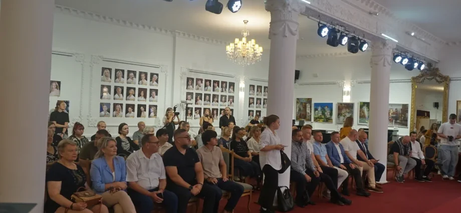Глядачі сидять у залі театру з колонами, люстрою та портретами на стіні